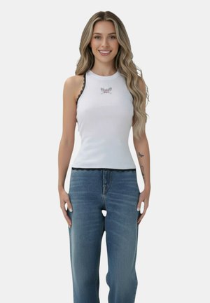 Donna sorridente con capelli biondi lunghi che indossa una canottiera bianca con rifiniture in pizzo nero e jeans blu, in piedi davanti a uno sfondo semplice.