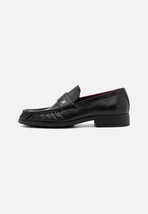 Mocasines de cuero negro con una textura suave, que presentan una correa decorativa en la parte frontal y un tacón ligeramente elevado, diseñados para comodidad y estilo.