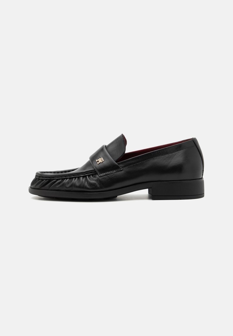 Mocassins en cuir noir au toucher lisse, avec une sangle décorative à l'avant et un talon légèrement surélevé, conçus pour le confort et le style.