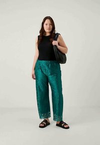 Zöldeskék csipkés nadrág, levélmintával, fekete ujjatlan toppal és fekete szandállal párosítva. A modellen fekete válltáska van.