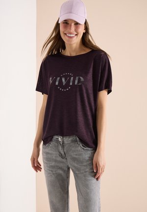 Kobieta uśmiechająca się, ubrana w jasnofioletową czapkę, ciemnofioletową koszulę z napisem "VIVID DREAMS" oraz jasnoszare dżinsy, stojąca na dwukolorowym tle.