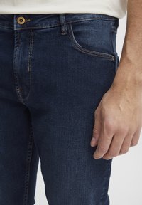 Jeans de mezclilla azul oscuro con un corte ajustado, que presentan herrajes dorados, costuras visibles y un clásico diseño de cinco bolsillos. La textura es suave.