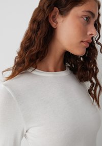 Weißes Baumwollhemd mit langen Ärmeln und rundem Ausschnitt, das über eine glatte Textur und subtile Nähte an den Schultern verfügt.