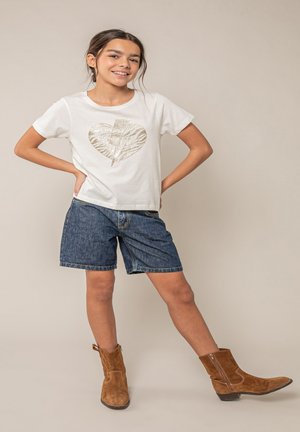 Fille souriante posant les mains sur les hanches, portant un t-shirt blanc avec un dessin de cœur et d'éclair, un short en denim et des bottes en suède marron.