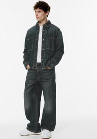 Donkerdenim jack met twee voorzakken, gecombineerd met losse donkere spijkerbroeken en witte sneakers. Model staat tegen een witte achtergrond.