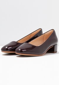 Talons en cuir verni bordeaux avec une pointe aigüe, un petit talon carré et une texture lisse. L'intérieur présente une semelle beige.