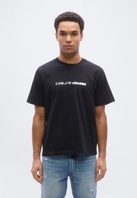 Ung mand set forfra iført en sort Calvin Klein t-shirt og slidte lyseblå denimshorts mod en ensfarvet hvid baggrund.