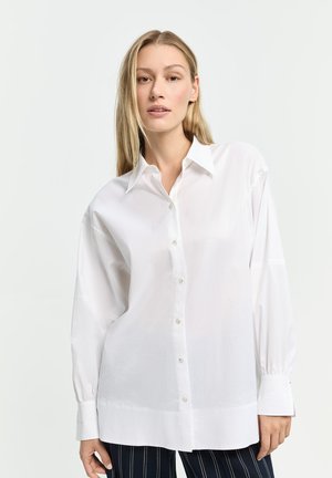 Frau trägt übergroßes weißes Hemd mit Knöpfen und schwarze Hose mit weißen Streifen, steht vor schlichtem hellem Hintergrund.