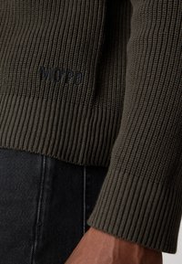 Suéter de punto verde oliva con textura acanalada y el logo "MOPD". El suéter presenta una silueta ajustada y mangas largas, combinado con jeans negros.