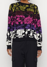 Pull avec motif floral horizontal en olive, marron, rose, violet et blanc sur fond noir, porté avec un pantalon noir.