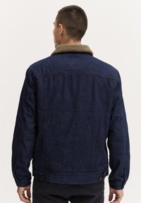 Donkerblauwe denimjas met een bruine fleece kraag, knoopsluiting bij de manchetten en een rechte pasvorm, met zichtbare stiksels op de achterkant.