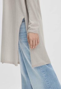 Lichtgrijze longsleeve top met zijsplitten, gecombineerd met wijde blauwe spijkerbroek. De stof lijkt zacht met een gladde textuur.