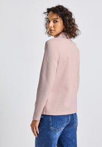 Un haut à manches longues rose pâle avec un col haut, une texture douce et un ourlet arrondi, associé à un jean bleu et une coupe décontractée.