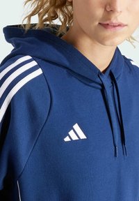 Marineblaue Kapuzenjacke aus weichem Stoff, mit weißen Streifen an den Ärmeln und einem weißen Logo auf der Brust. Verstellbare Kapuze mit Kordelzug.