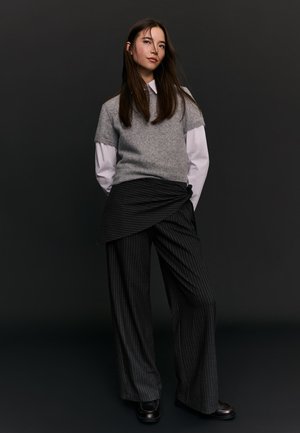 Mlada ženska z dolgimi rjavimi lasmi, oblečena v sivi pulover z kratkimi rokavi preko bele srajce, črne široke hlače s črtami in črne čevlje.