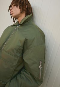 Giacca bomber verde oliva con colletto alto, chiusura frontale con zip e tasca con zip sulla manica; realizzata in materiale liscio e leggero.