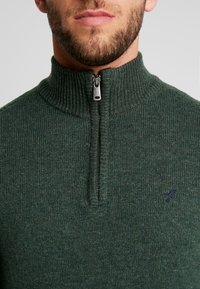 Uomo che indossa un maglione lavorato a maglia verde con colletto a mezza zip e un piccolo logo scuro sul lato sinistro del petto, visibile dalle spalle alle labbra.