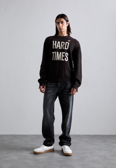Schwarzer gestrickter Pullover mit dem Aufdruck "HARD TIMES", verwaschene Details. Kombiniert mit dunkelblauen Jeans und weißen Sneakers mit hellbrauner Sohle.
