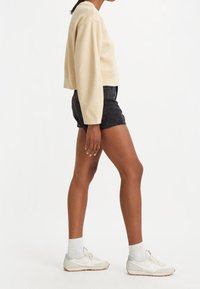 Mujer de pie de perfil, vistiendo un sudadera corta de manga larga beige, pantalones cortos de mezclilla negros, calcetines blancos y zapatillas Nike blancas sobre fondo blanco.