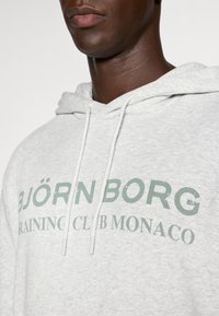 Ανοιχτό γκρίζο φούτερ από μείγμα βαμβακιού, με κορδόνια και μπροστινή εκτύπωση που αναγράφει "BJÖRN BORG RAINING CLUB MONACO" σε πράσινο χρώμα.