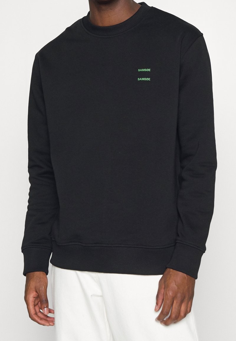 Sweat-shirt noir à manches longues et col rond avec un petit texte vert « SAMSOE » imprimé deux fois sur la poitrine gauche, porté avec un pantalon blanc.