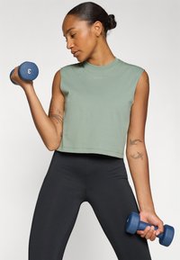 Green sleeveless crop top with a smooth texture. Black fitted leggings. Holding two blue dumbbells, one marked with the number 3.