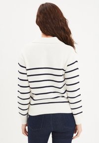 Maglione bianco con strisce orizzontali blu navy, orlo e polsini a costine, design con colletto e vestibilità rilassata, abbinato a jeans scuri.