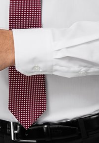 Witte overhemdd met knoopmanchetten, gecombineerd met een bordeauxrode das met een ruitpatroon. Zwarte riem zichtbaar op de taille.
