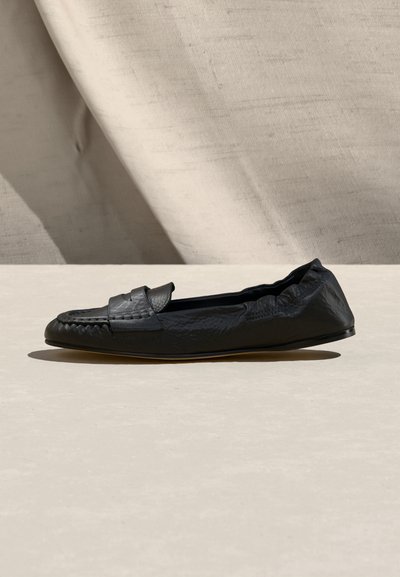 Μαύρο δερμάτινο loafers χωρίς κορδόνια με υφή, επίπεδη σόλα και λεπτομέρεια λουριού penny, τοποθετημένο σε μπεζ επιφάνεια με υφασμάτινο φόντο.