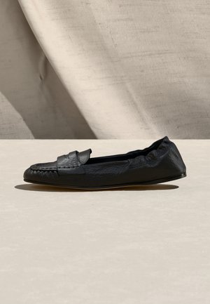 Must nahast libisev loafer, millel on tekstuuriga viimistlus, lame tald ja mündi rihma detail, asetatud beežile pinnale kangataustal.
