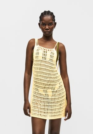 Γυναίκα με πλεξούδες φοράει αμάνικο, κίτρινο πλεκτό φόρεμα πάνω από ελαι πράσινη βάση, μπροστά σε ένα απλό φωτεινό φόντο.