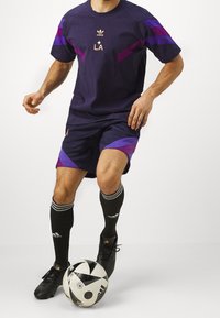 Adidas purple urheiluasu, joka sisältää lyhythihaisen paitaan LA-logolla ja tummat shortsit, jotka ovat kontrastiväriset. Mustat sukat ja kengät sekä musta-valkoinen jalkapallo.