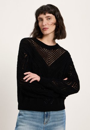 Frau mit dunklem Bob-Haar, die einen schwarzen, offen gestrickten Pullover und hellblaue Jeans trägt, steht mit verschränkten Armen vor einem einfarbigen Hintergrund.