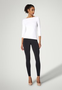 Weißes Top mit drei Viertelärmeln kombiniert mit schwarzen Leggings und nudefarbenen High Heels, das ein figurbetontes Design mit glatter Textur präsentiert.