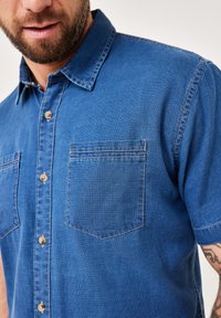 Kurzarm-Denim-Hemd in Mittelblau, mit einer Knopfverschlussleiste und zwei Brusttaschen mit kontrastierenden Nähdetails.