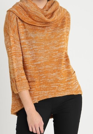Femme portant un pull en tricot marbré orange et blanc, ample, avec des manches 3/4 et un large col bénitier, associé à un pantalon noir.