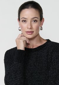 Črna pletena majica z teksturiranim vzorcem, z ribičastimi manšetnimi rokavi in ovratnikom. Model nosi srebrne obroče in nevtralen videz ličil.