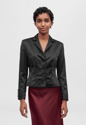 Une femme porte un blazer noir en satin cintré à boutons et une jupe bordeaux sur un fond clair uni.