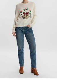 Kremowy, dziergany sweter z dekoracyjnym wzorem dzwonka i liści ostrokrzewu, zestawiony z jasnoniebieskimi jeansami i brązowymi botkami na obcasie.