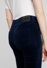 Marinblå kordflöjelbyxor med en svart Lee-logotyp på baksidan. Har svängda fickor och en figursydd design. Slät textur.