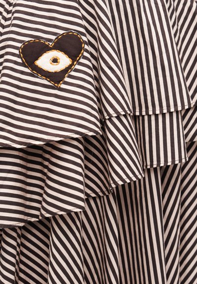 Υφασμα με ασπρόμαυρες ρίγες, διακοσμημένο με στρώσεις βολάν και ένα μικρό μαύρο μπαλώμα σε σχήμα καρδιάς που φέρει κεντημένο λευκό μάτι με πορτοκαλί ραφές.