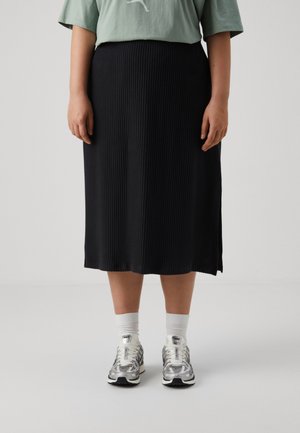Osoba ubrana w czarną dzianinową midi spódnicę, białe skarpetki, srebrne buty sportowe Nike i jasnooliwkową koszulkę Puma stoi na tle jednolitego tła.