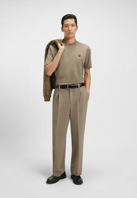 T-shirt beige a maniche corte con logo, pantaloni beige su misura, cintura nera e scarpe nere, con una giacca marrone poggiata sulla spalla.