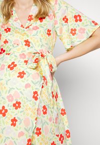 Vestido envolvente floral em tecido suave e leve. Base amarelo pálido com flores vermelhas e rosas, folhas verdes e detalhe de cintura amarrada. Mangas curtas.