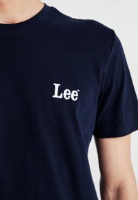 Mørkeblå bomull T-skjorte med en hvit brodert "Lee"-logo på venstre brystområde; rund hals og korte ermer.