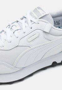 Chaussure de sport blanche avec un mélange de cuir lisse et de mesh, dotée d'un bout arrondi, d'un laçage à l'avant et de détails de marque.