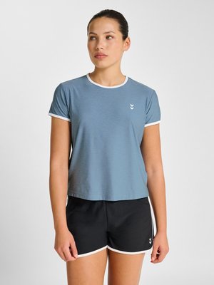 Vrouw draagt een blauw T-shirt met korte mouwen en zwarte sportshorts met witte rand, staand tegen een effen lichte achtergrond.