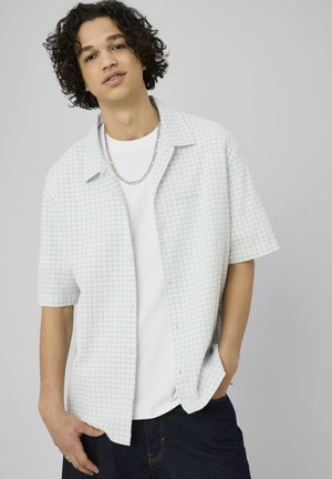 Jeune homme aux cheveux bouclés portant une chemise à manches courtes à carreaux claire sur un tee-shirt blanc, un jean foncé et un collier chaîne en argent.
