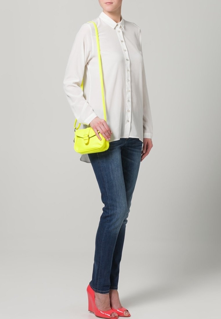 Camicia bianca a bottoni, jeans skinny blu, décolleté rosa a punta aperta e una piccola borsa crossbody gialla neon. Tessuto morbido e vestibilità casual.