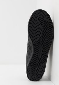 Musta Adidas-tennarien pohja, jossa on sik-sak ja aaltoileva renkaat, asetettuna pystysuuntaan valkoista nurkkaseinää vasten.